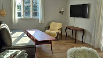 A living room with a sofa, a wooden table, a fluffy armchair, a television, and a window with curtains.