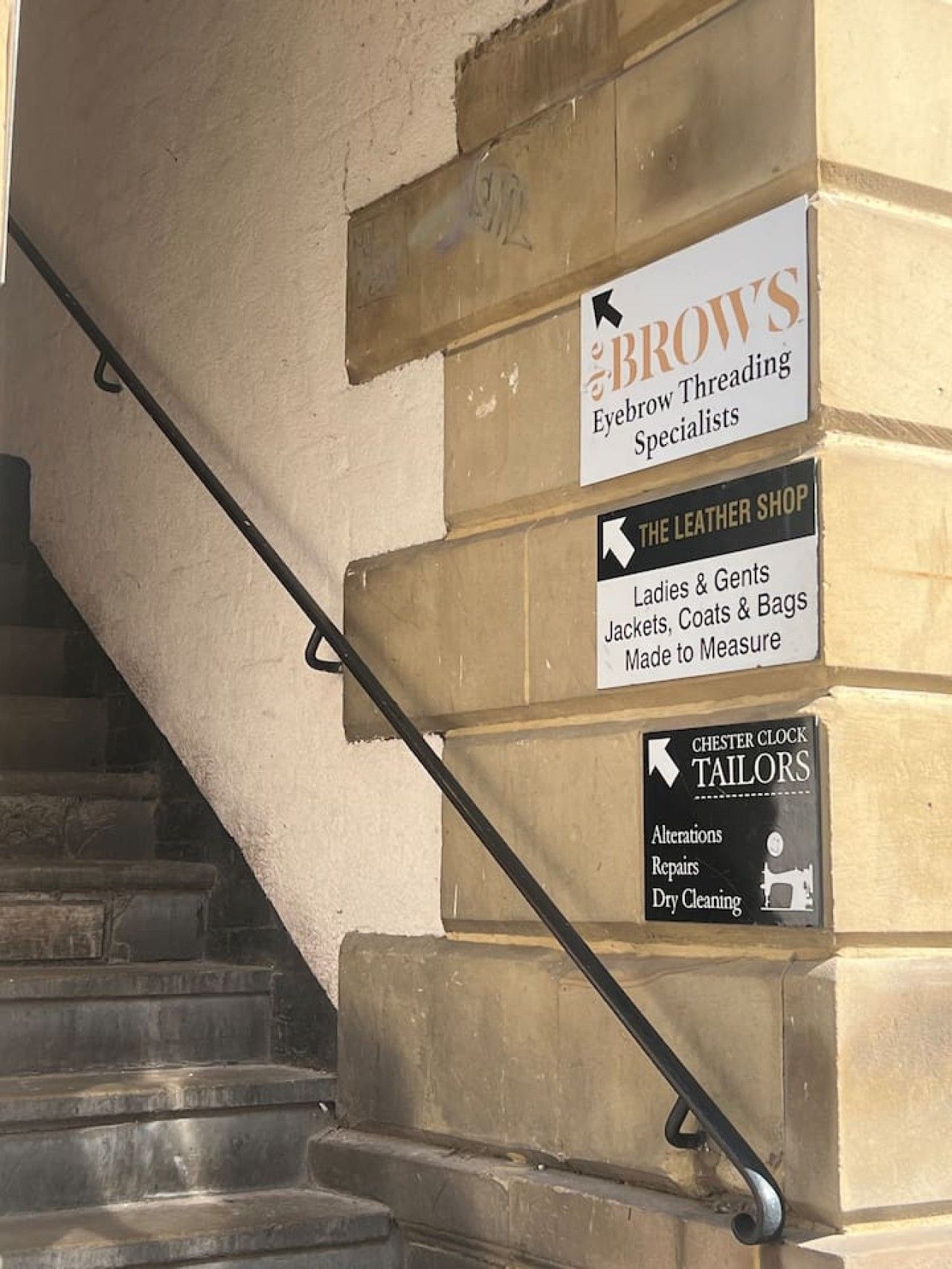 Eine Treppe mit Handläufen und Schildern für Brow's Eyebrow Threading Specialists, The Leather Shop und Chester Clock Tailors.