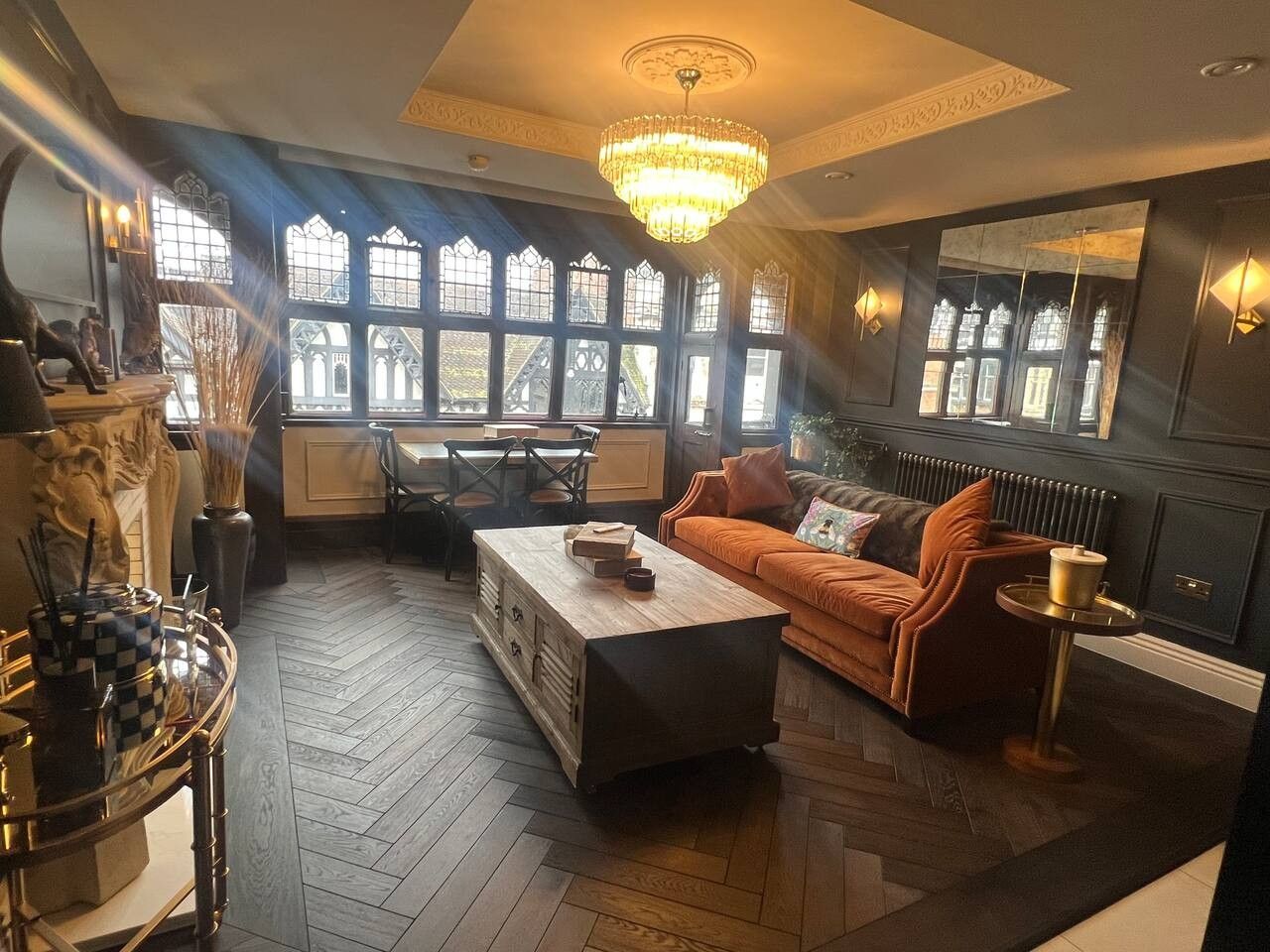 Ein Zimmer mit Holztisch, einer Couch, einem Kronleuchter und großen Fenstern mit verzierten Glasscheiben.