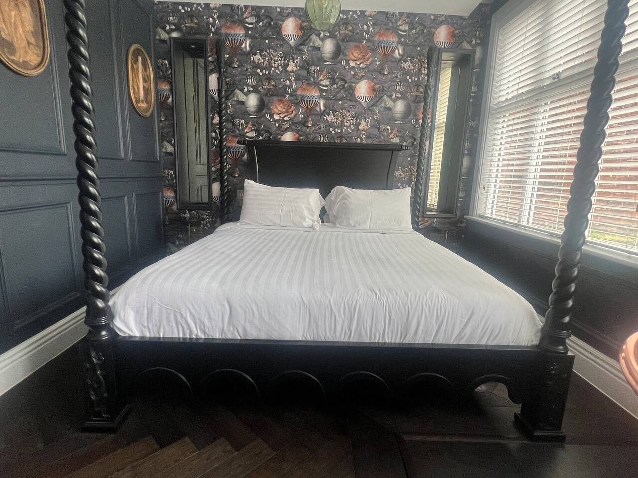 A four-poster bed with a dark frame and white bedding in a room with patterned wallpaper and a window with blinds.