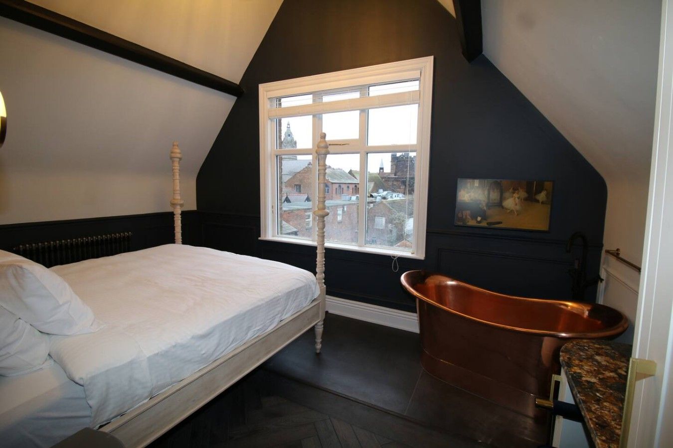 A bedroom with a bed, a window with a view of buildings, a copper bathtub, and a painting on the wall.