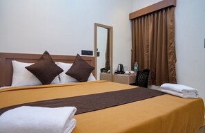 A hotel room with a bed, pillows, a bedside table with a kettle, a chair, and a window with curtains.