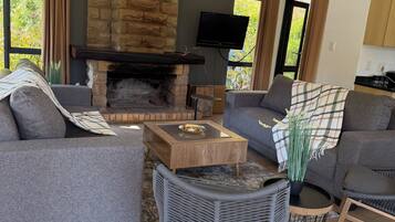 A living room with a stone fireplace, grey sofas, a television, and a view of greenery outside.