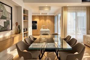 A modern living room with a glass-top dining table surrounded by cushioned chairs, a kitchen area with built-in ovens, and a large window with sheer curtains. - Genteel Home Reyes Catolicos Sevilla (Seville)