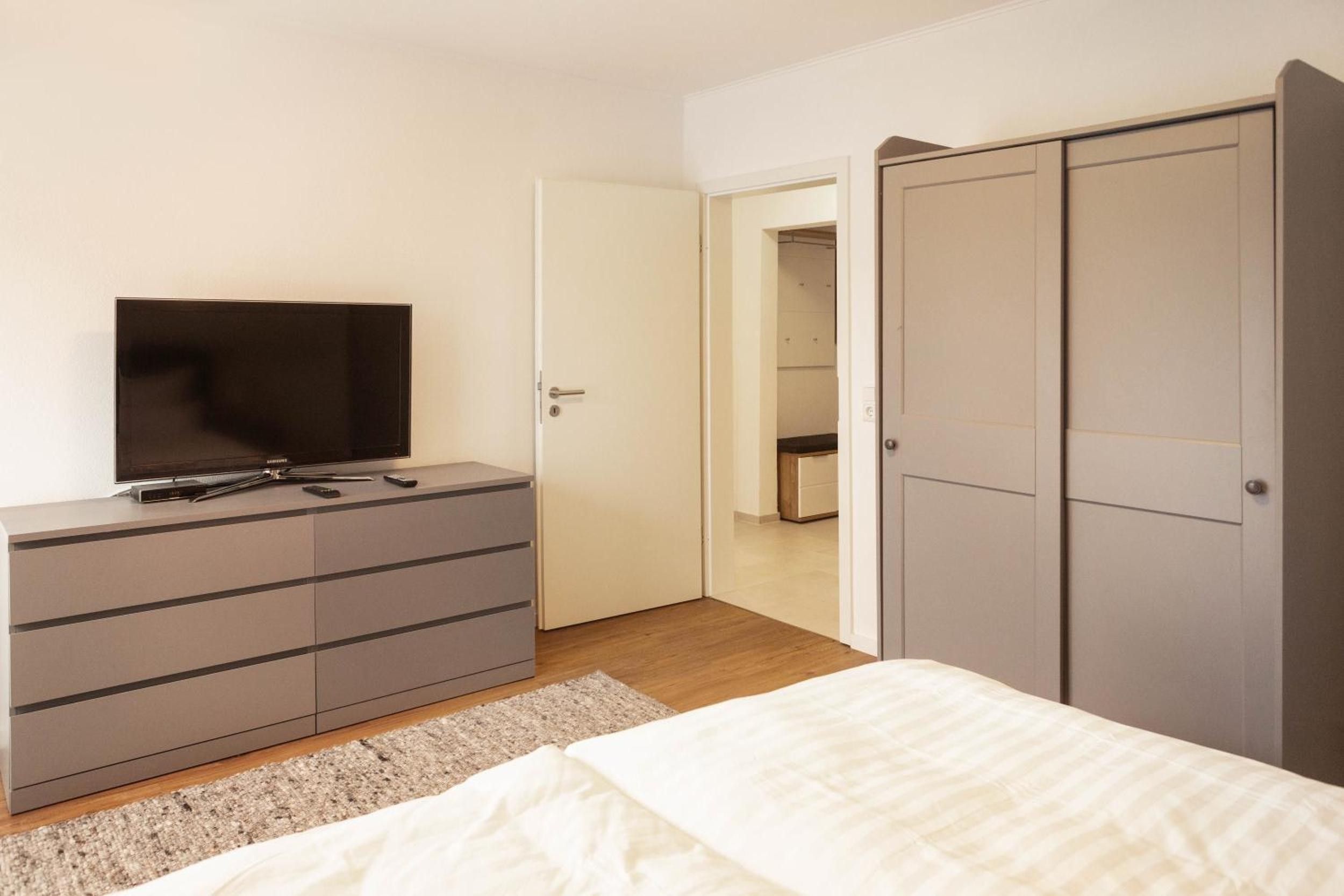 Ein Schlafzimmer mit einem Bett, einem Fernseher auf einer Kommode, einem Kleiderschrank und einer Tür zu einem anderen Raum.