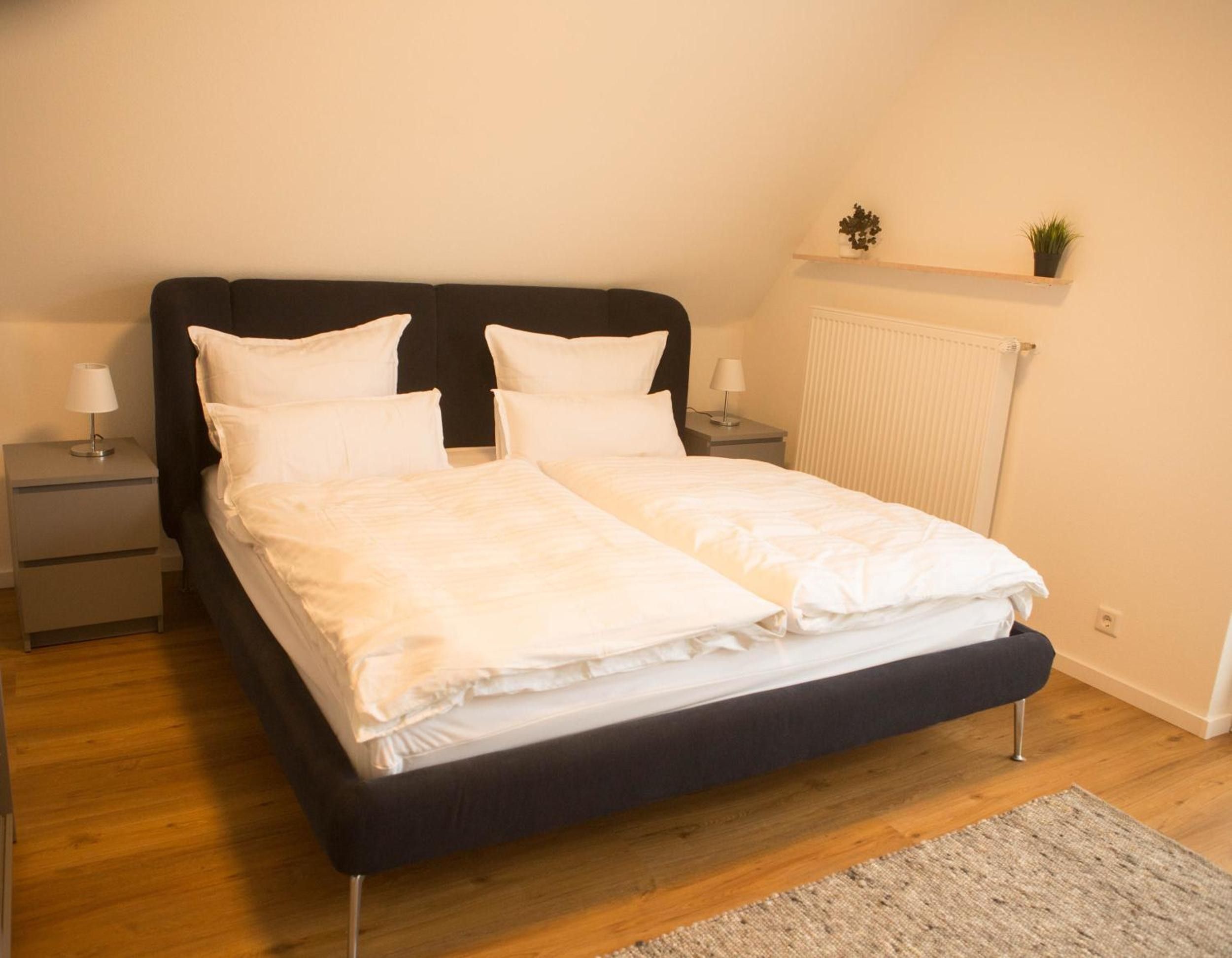 Ein Doppelbett mit weißer Bettwäsche, ein dunkles Kopfteil, zwei Nachttische mit Lampen, eine Heizung und eine kleine Ablage mit Pflanzen.