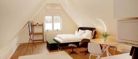 Un dormitorio con cama, mesita de noche, silla y escritorio con otra silla. Se ve una ventana con vista a árboles.
