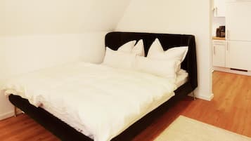 Un lit simple avec une tête de lit noire et du linge de lit blanc, dans une pièce au plancher en bois et aux murs blancs.