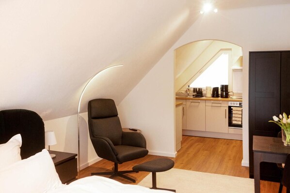 Een moderne slaapkamer met een enkel bed, een donkere fauteuil en een kleine tafel. In de achtergrond is een keuken te zien met een eethoek.