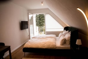 Een slaapkamer met een bed, een aan de muur bevestigde televisie, een raam met uitzicht op bomen en een lamp op een nachtkastje.
