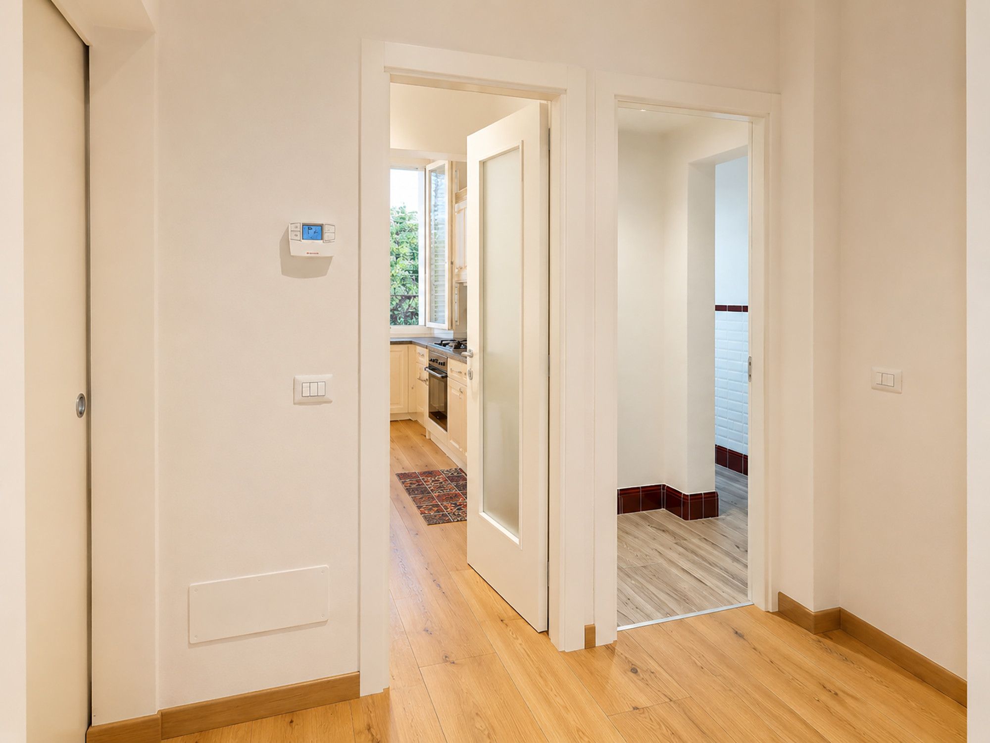 A hallway with wooden flooring, white walls, and a view into a kitchen area with wooden cabinets and a window.
