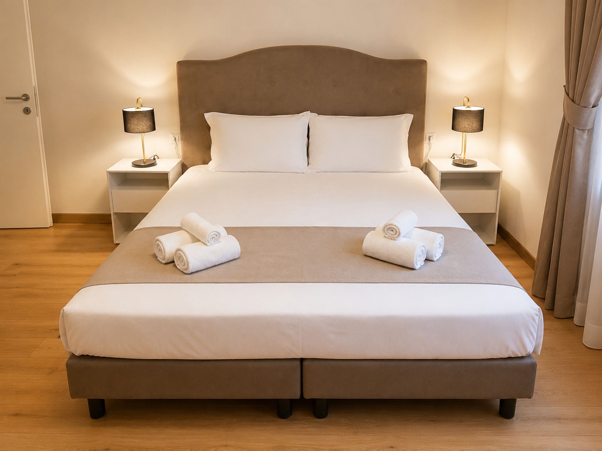 A double bed with white linens, two bedside tables with lamps, and a pair of rolled towels on the bed.