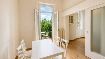 A room with a white table and chairs, a radiator, a door with frosted glass, and a view of greenery outside.