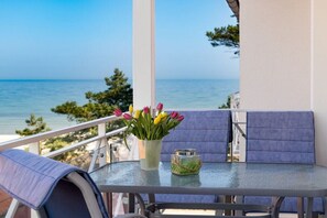 Un balcón con mesa de vidrio, sillas con cojines azules, un florero con flores y vista al mar.