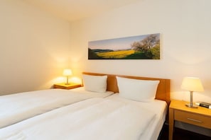 Una cama doble con ropa blanca, dos mesitas de noche con lámparas, y un cuadro de paisaje en la pared.