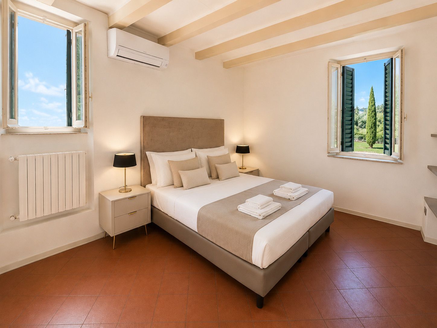 Quarto com cama, luminárias laterais, radiador e janelas com persianas.