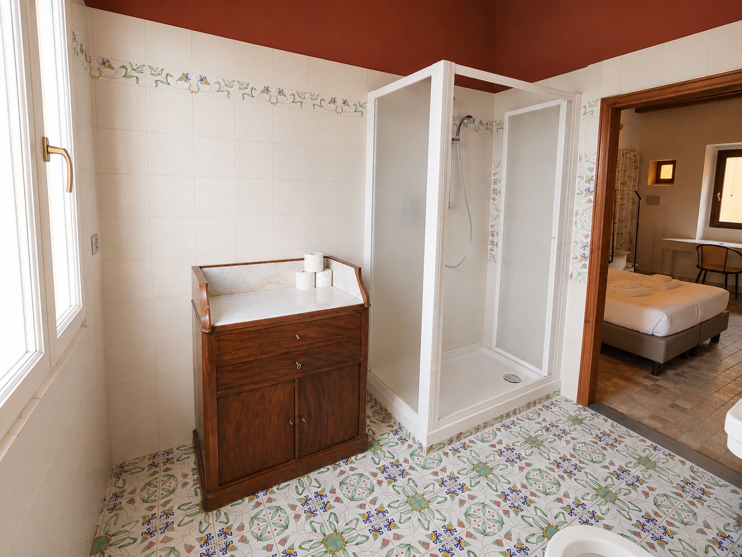 Un baño con un mueble de madera, cabina de ducha y una cama visible a través de una puerta abierta.