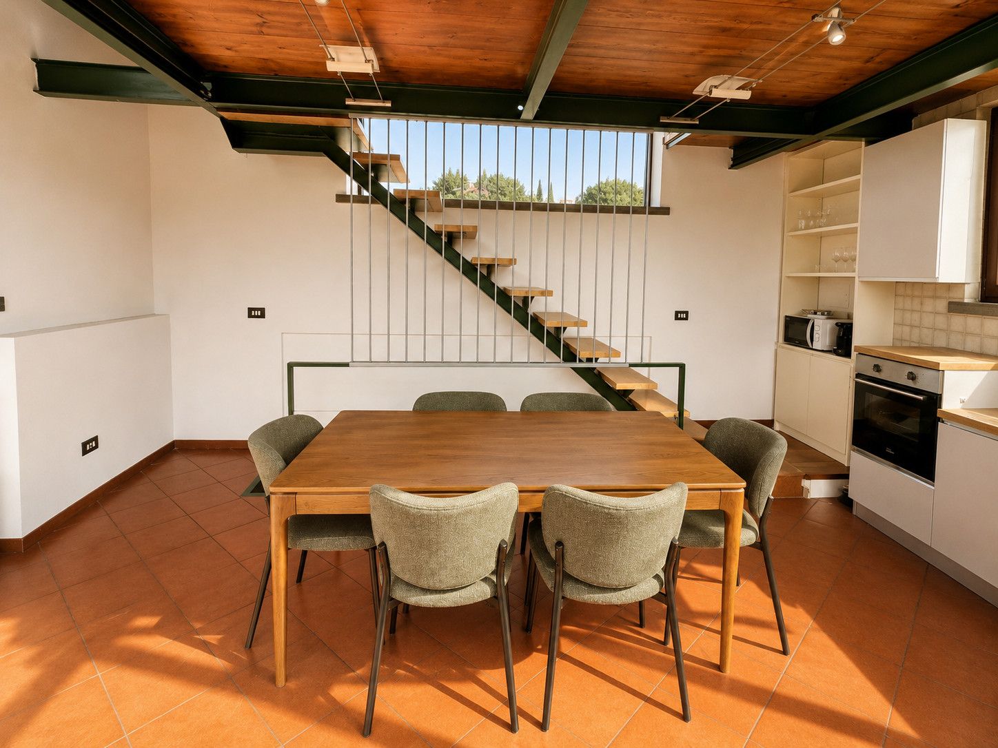 A modern kitchen with a wooden dining table and chairs, a staircase, and a view of the outdoors.