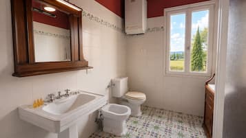 Ein Badezimmer mit einem weißen Waschbecken, einer Toilette, einem Bidet, einem Spiegel und einem Fenster mit Blick auf Grünflächen.