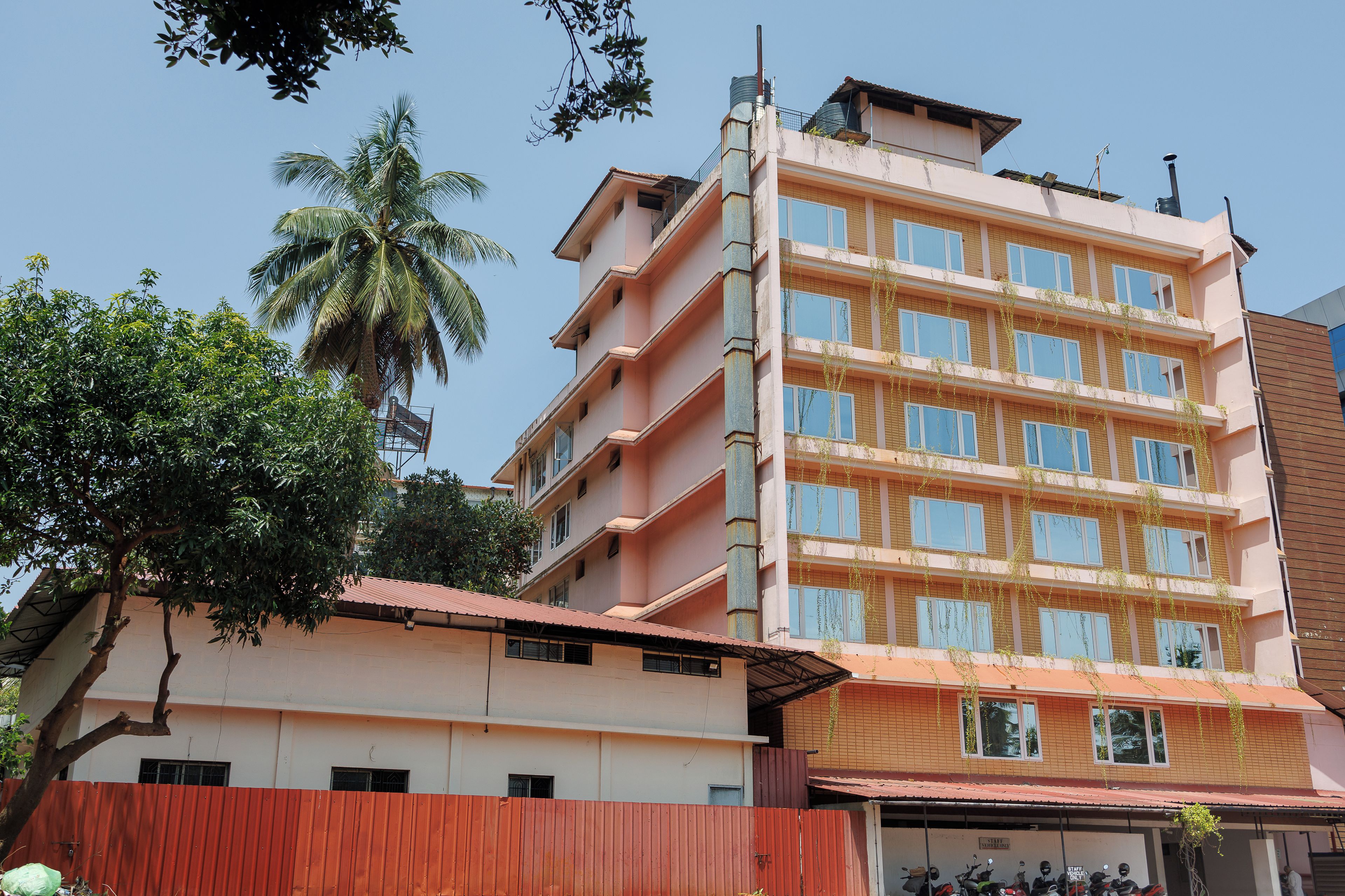 Un edificio de varios pisos con una cerca roja al frente, una palmera y un cielo despejado.