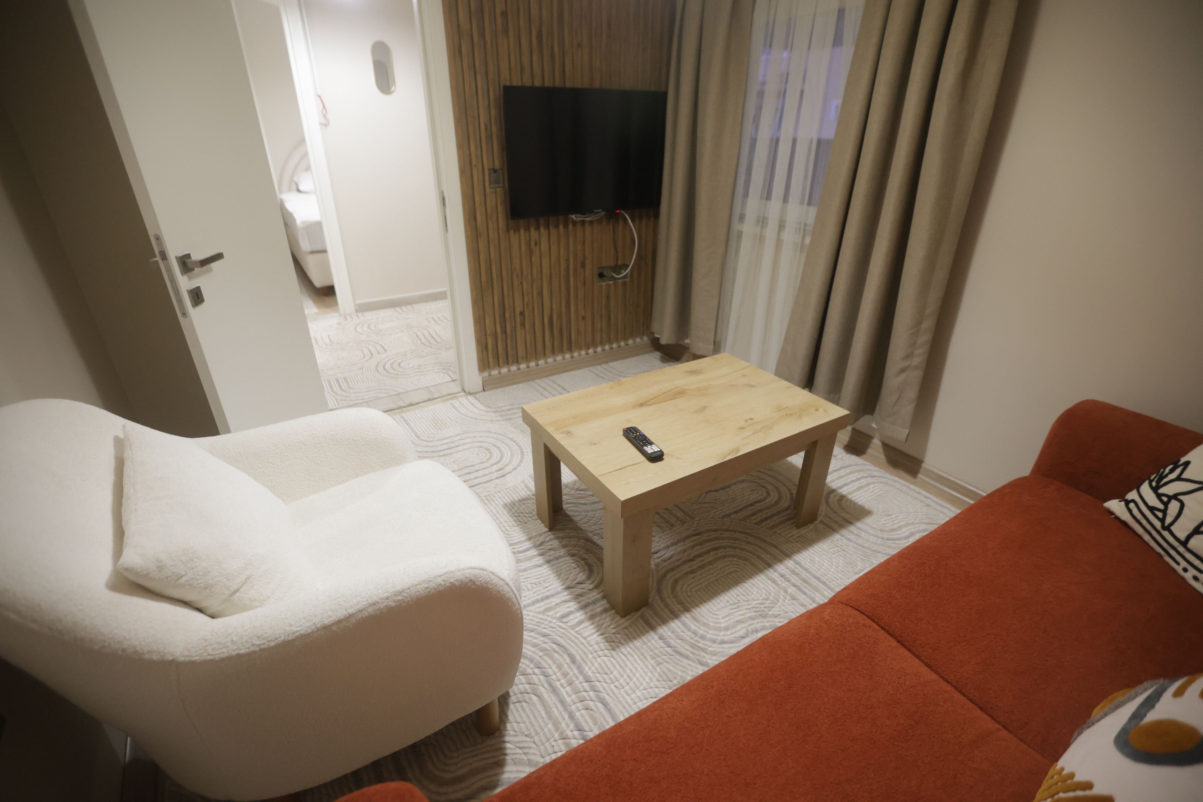 Ett rum med en vit fåtölj, en orange soffa, ett träbord och en tv som är monterad på en vägg av trä.