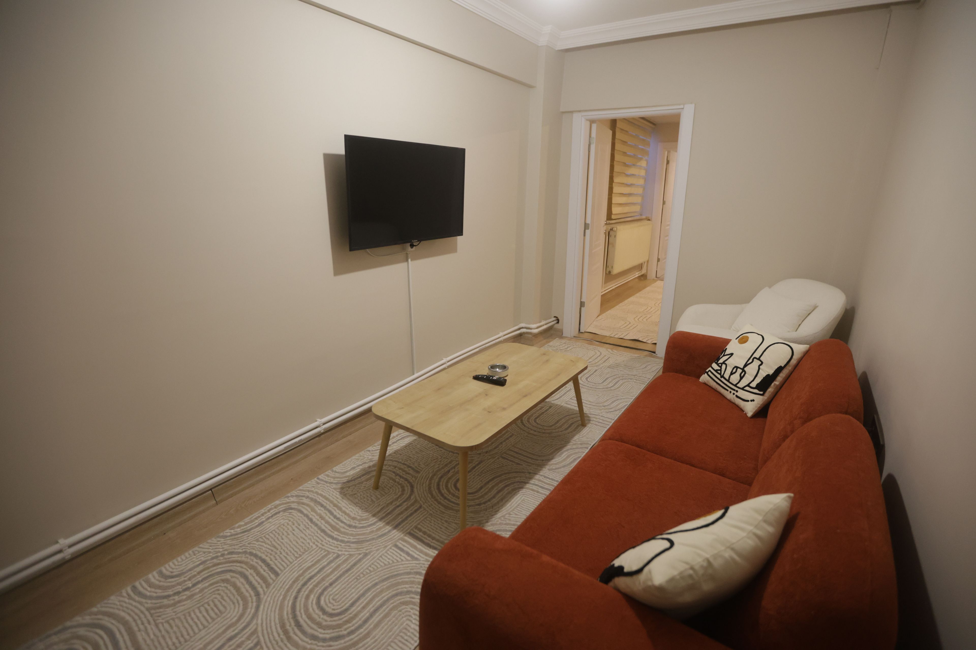 A living room with a flat-screen TV mounted on the wall, a wooden coffee table, a red sofa with a patterned cushion, and a patterned rug on the floor.