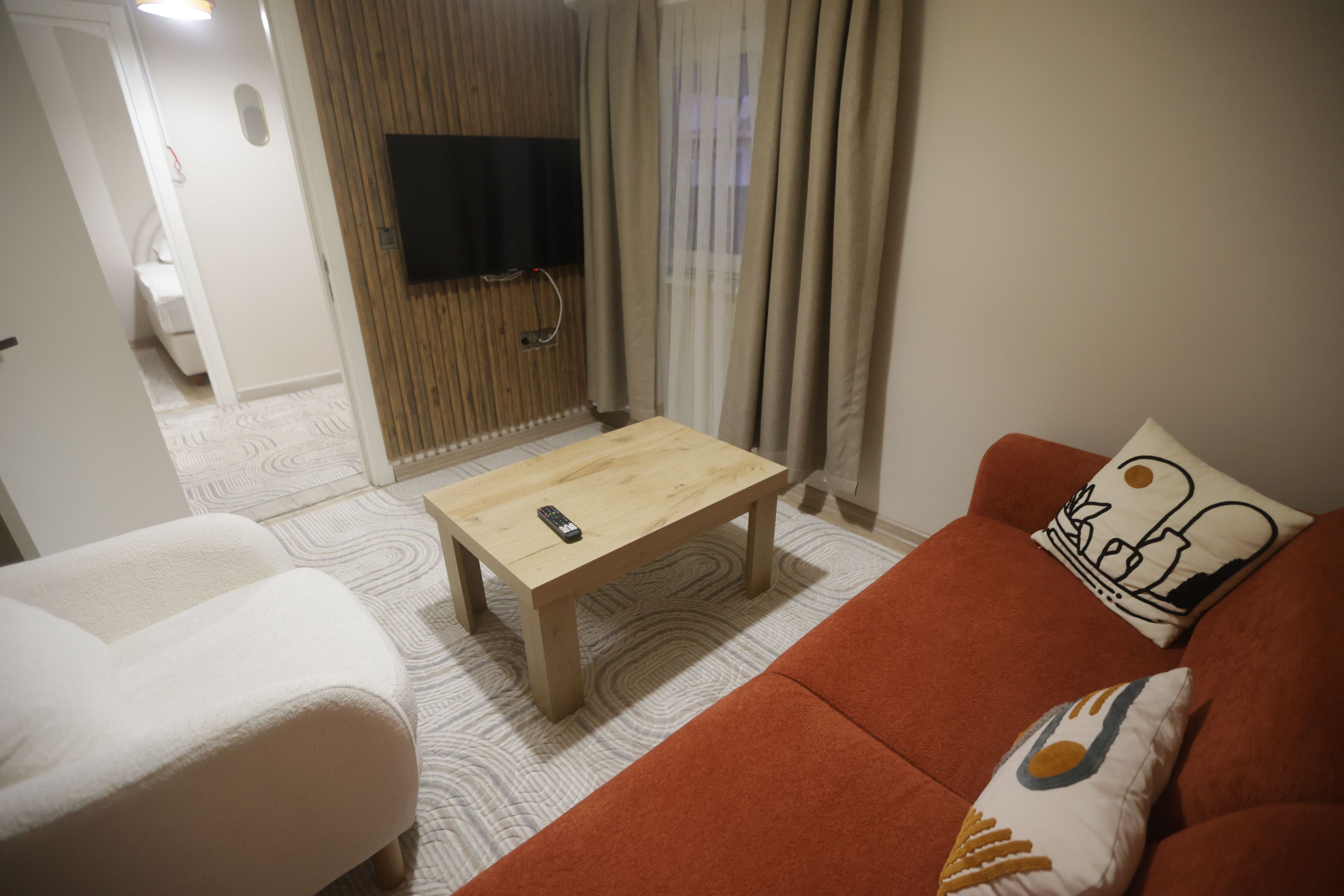 A living room with a wooden coffee table, a sofa with a cushion, a television mounted on a wooden panel, and a patterned rug.