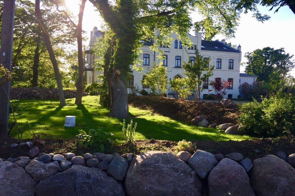 A large, white building with multiple windows, surrounded by a well-maintained garden with trees and a stone wall.
