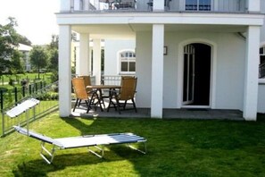 Ein überdachter Außenbereich mit Tisch, Stühlen und einer Lounge-Liege, sowie ein weißes Gebäude mit einer Tür.
