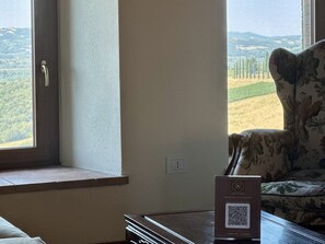A room with a window overlooking a landscape, a floral-patterned chair, and a QR code on a table. - Camigliano Wine Estate - Oliviera (Camigliano)
