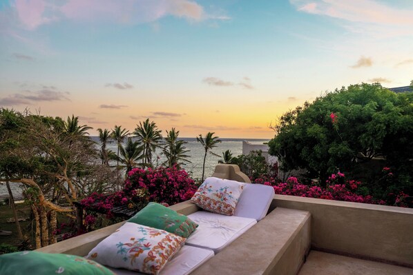 A lounge area with a view of the sea, featuring a sofa with patterned cushions and a backdrop of lush greenery and pink flowers.