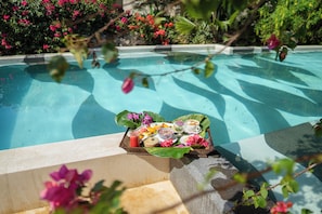 Escena junto a la piscina con una bandeja de comida y bebidas, rodeada de exuberante vegetación y flores.