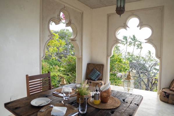Mesa de comedor dispuesta con platos, cubiertos y una copa, con vista a follaje a través de ventanales arqueados.