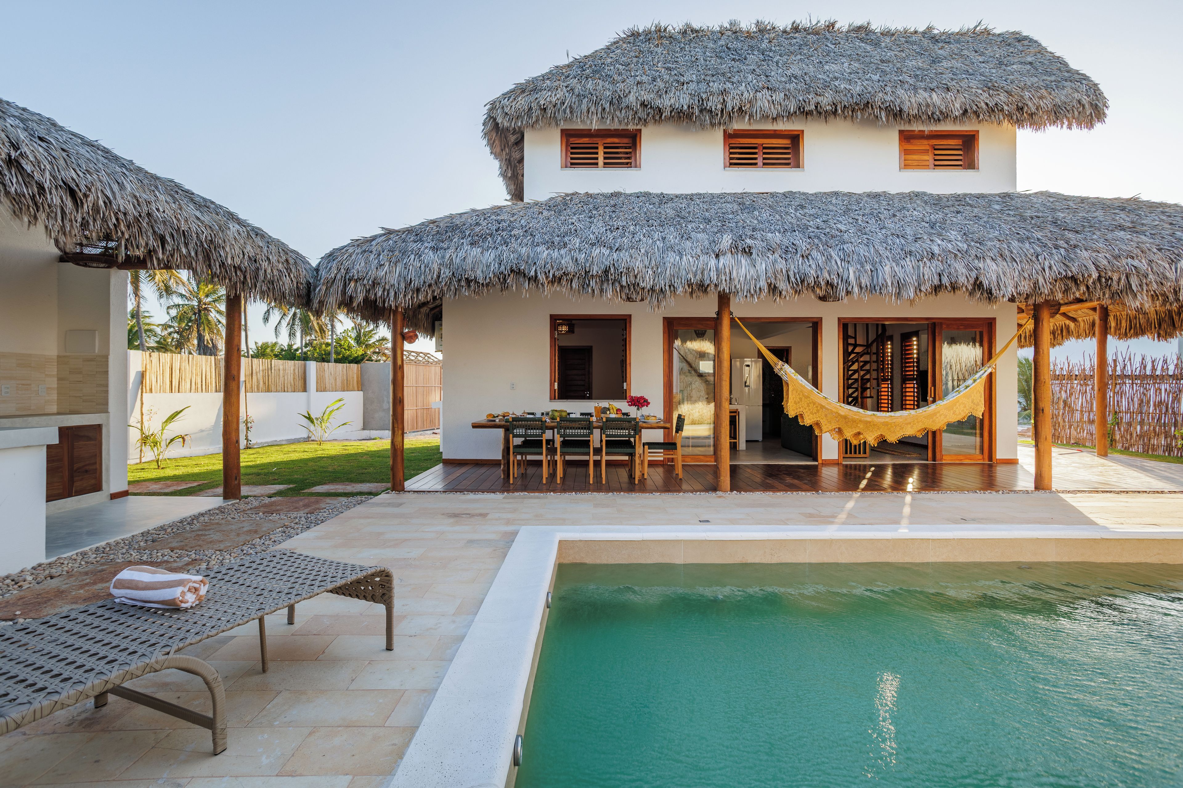 Un espace aménagé au bord de la piscine, avec une structure à toit de chaume, une table à manger et un hamac.
