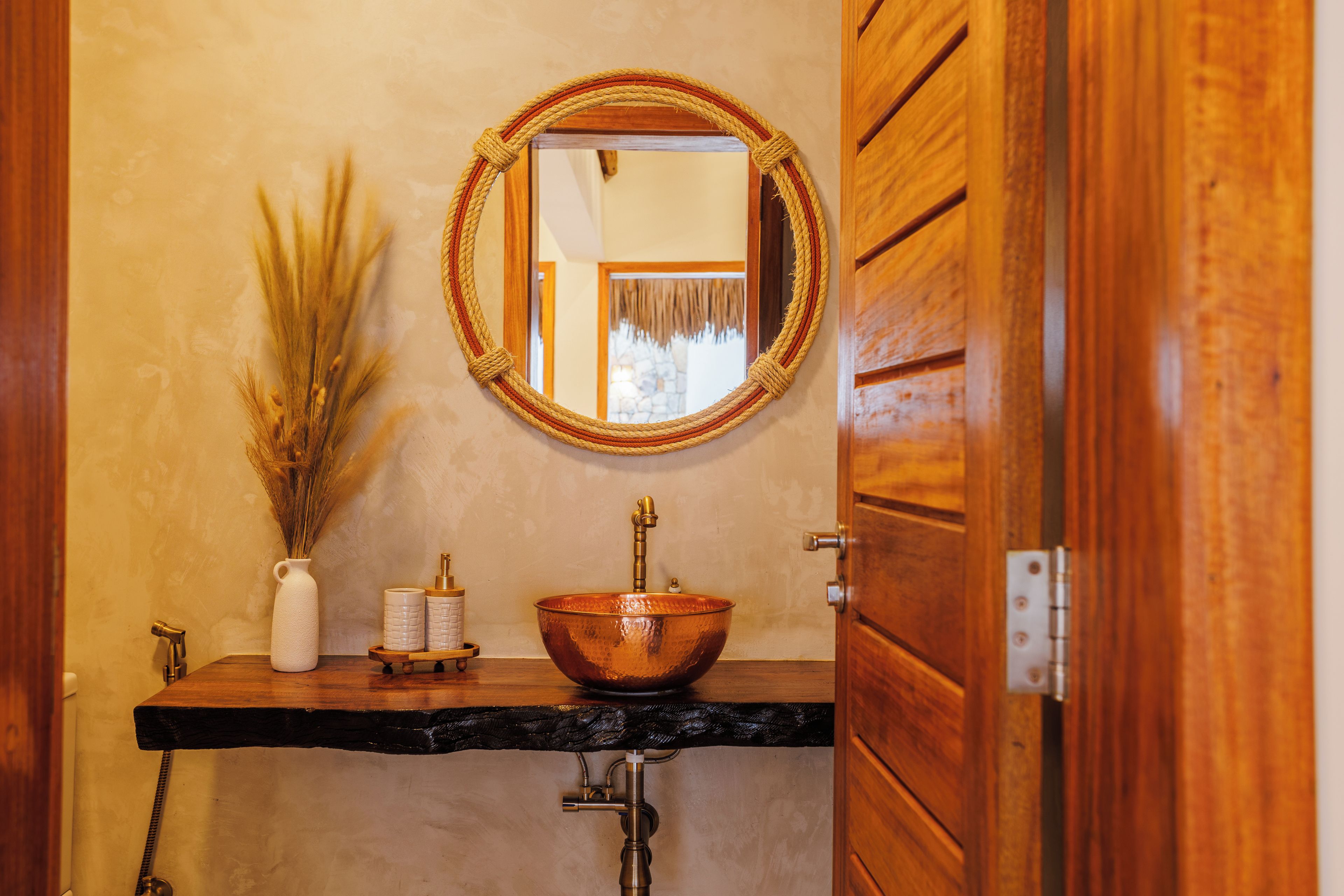 A bathroom with a wooden vanity, a copper basin, a white vase with dried plants, and a mirror on the wall.