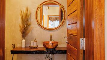 A bathroom with a wooden vanity, a copper basin, a white vase with dried plants, and a mirror on the wall.
