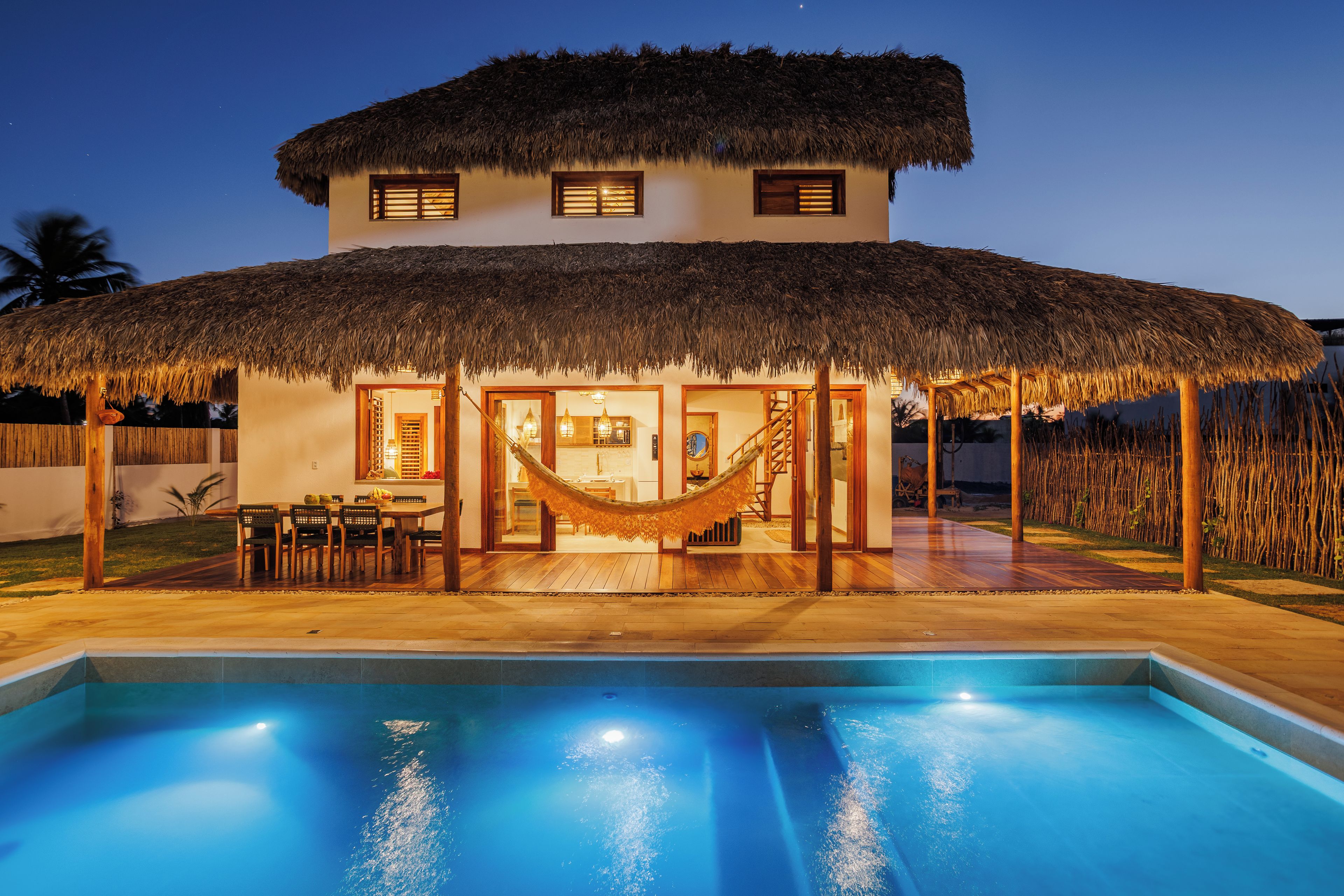 Une maison avec un toit de chaume, une piscine et un coin repas aménagé avec des meubles en bois.