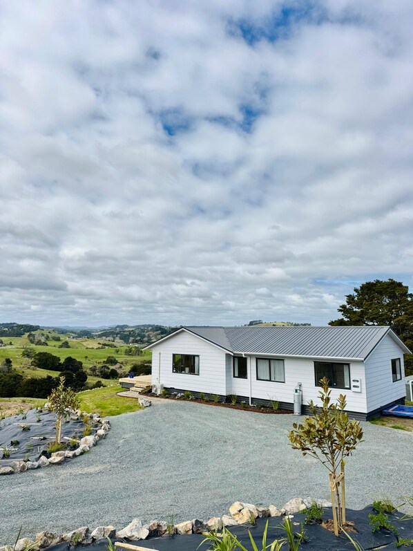 A single-story white house with a grey roof, surrounded by a gravel driveway and a small garden with young trees. - Escape the city and slow down at 'The Ranch', 1.5 hours North of Auckland. (Paparoa)