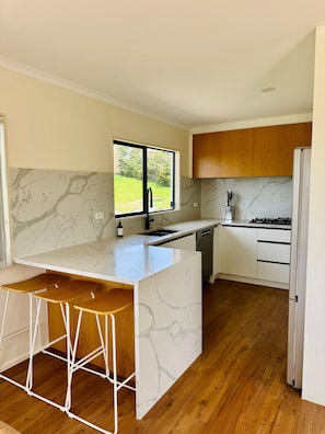 A modern kitchen with a marble countertop, wooden bar stools, a built-in sink, and a view of a green landscape through a window. - Escape the city and slow down at 'The Ranch', 1.5 hours North of Auckland. (Paparoa)