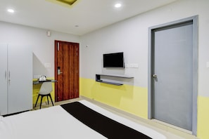 Una habitación de hotel con cama, escritorio con silla, televisor montado en la pared y una puerta.