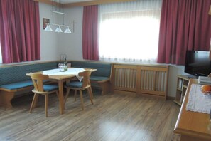 A room with a dining table, chairs, a bench, a radiator, a television, and curtains. - Holiday apartment with 2 bedrooms (Nauders)