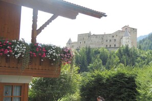 A building with a balcony adorned with flowers, a wooden structure, and a castle in the background. - Holiday apartment with 2 bedrooms (Nauders)