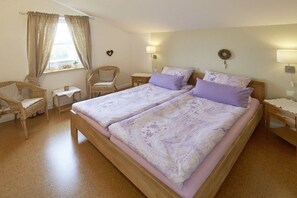 A double bed with a floral bedspread, two bedside tables, a nightstand, a wicker chair, a small table, and a window with curtains. - Holiday home with 2 bedrooms (Wackerballig)