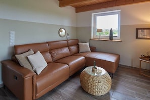 Ein braunes, ledernes Ecksofa mit einem runden Gewebecouchtisch und einem kleinen Beistelltisch, ein Fenster mit Blick und ein wandmontiertes Spiegel.