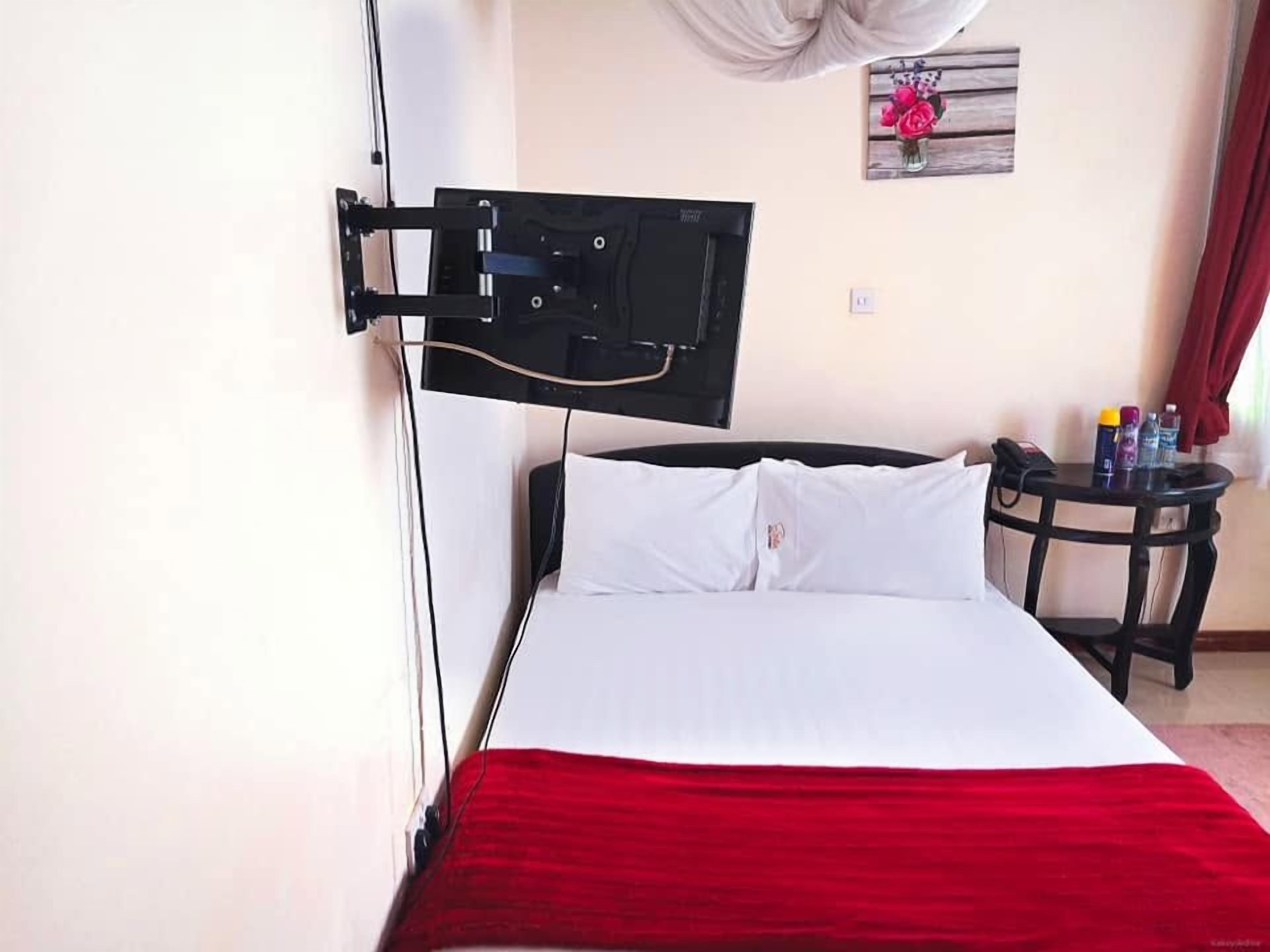 A bed with white bedding and a red accent, a mounted television on the wall, a small round table with a chair, and a wall-mounted picture.