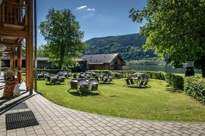 A lakeside outdoor seating area with chairs and tables, a wooden building, and a view of a lake and hills. - Holiday apartment with 1 bedroom (Steindorf)