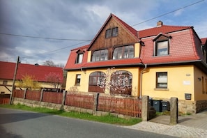 A two-story house with a red roof and yellow walls, a wooden fence, and a paved driveway. - Holiday apartment with 1 bedroom (Rathewalde)