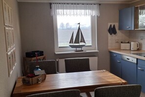 A compact kitchen with a dining table, chairs, a window with a sailboat decoration, and a view of the water. - Holiday apartment with 2 bedrooms (Sehestedt)