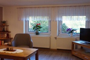 A living room with a wooden table, a chair, a television on a wooden stand, and two windows with curtains. - Holiday apartment with 2 bedrooms (Bad Schandau Stadtteil Prossen)