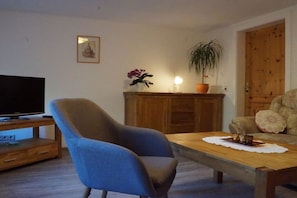 A living room with a television on a wooden stand, a blue armchair, a wooden coffee table, a potted plant, and a wooden cabinet. - Holiday apartment with 2 bedrooms (Bad Schandau Stadtteil Prossen)